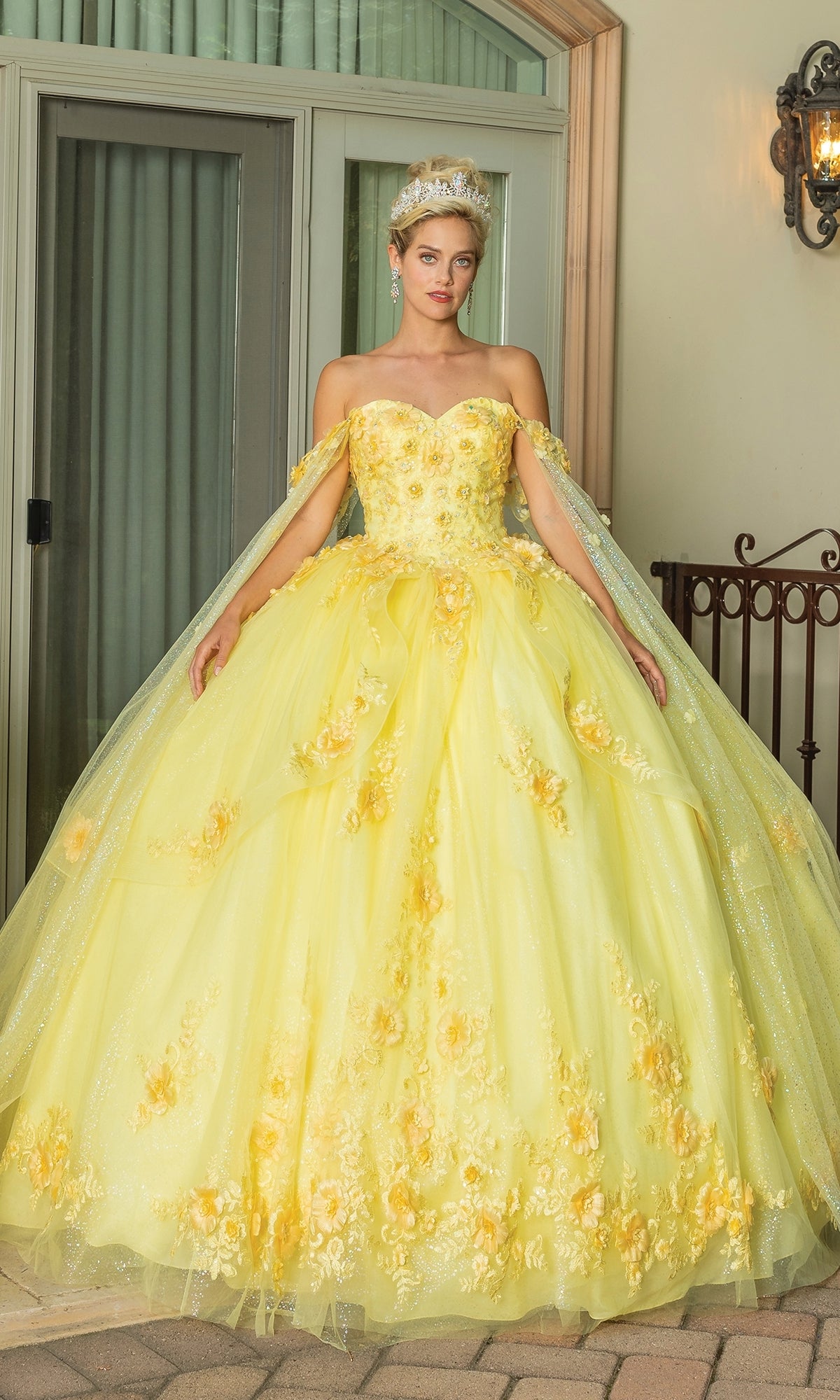 A woman stands by a window and railing wearing Quinceanera Dress 1788 By Dancing Queen, a yellow off-the-shoulder floral A-line ball gown with a sweetheart neckline and sheer cape.